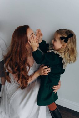 Beyaz bohem elbiseli güzel kızıl saçlı kadın küçük kızıyla temas kuruyor. Odada beyaz bir duvarın önünde. 