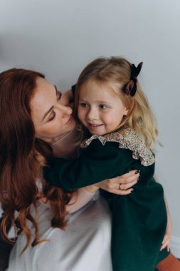 Beyaz bohem elbiseli güzel kızıl saçlı kadın küçük kızıyla temas kuruyor. Odada beyaz bir duvarın önünde. 