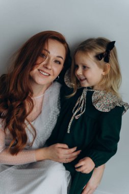 Beyaz bohem elbiseli güzel kızıl saçlı kadın küçük kızıyla temas kuruyor. Odada beyaz bir duvarın önünde. 