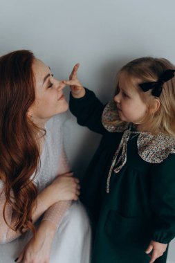 Beyaz bohem elbiseli güzel kızıl saçlı kadın küçük kızıyla temas kuruyor. Odada beyaz bir duvarın önünde. 