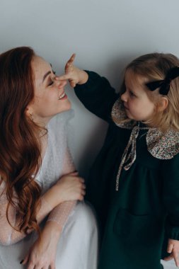 Beyaz bohem elbiseli güzel kızıl saçlı kadın küçük kızıyla temas kuruyor. Odada beyaz bir duvarın önünde. 