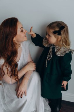 Beyaz bohem elbiseli güzel kızıl saçlı kadın küçük kızıyla temas kuruyor. Odada beyaz bir duvarın önünde. 