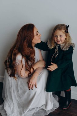 Beyaz bohem elbiseli güzel kızıl saçlı kadın küçük kızıyla temas kuruyor. Odada beyaz bir duvarın önünde. 