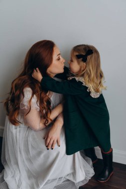 Beyaz bohem elbiseli güzel kızıl saçlı kadın küçük kızıyla temas kuruyor. Odada beyaz bir duvarın önünde. 