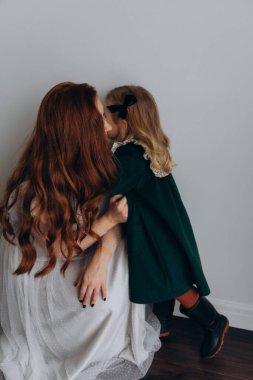 Beyaz bohem elbiseli güzel kızıl saçlı kadın küçük kızıyla temas kuruyor. Odada beyaz bir duvarın önünde. 