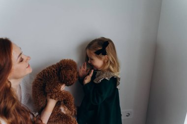 Beyaz bohem elbiseli güzel kızıl saçlı kadın bir köpek tutuyor ve küçük kızına beyaz bir duvarın önünde onu gösteriyor. 