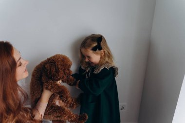 Beyaz bohem elbiseli güzel kızıl saçlı kadın bir köpek tutuyor ve küçük kızına beyaz bir duvarın önünde onu gösteriyor. 