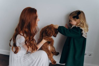 Beyaz bohem elbiseli güzel kızıl saçlı kadın bir köpek tutuyor ve küçük kızına beyaz bir duvarın önünde onu gösteriyor. 