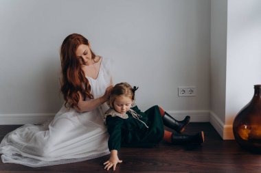 Beyaz bohem elbiseli güzel kızıl saçlı kadın küçük kızına örgü örüyor. Odada beyaz bir duvarın önünde. 