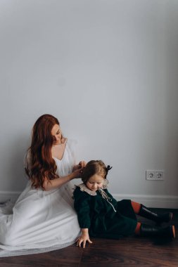 Beyaz bohem elbiseli güzel kızıl saçlı kadın küçük kızına örgü örüyor. Odada beyaz bir duvarın önünde. 