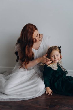 Beyaz bohem elbiseli güzel kızıl saçlı kadın küçük kızına örgü örüyor. Odada beyaz bir duvarın önünde. 