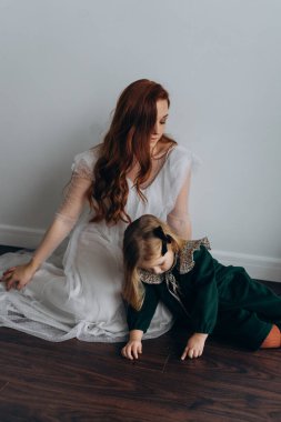 Beyaz bohem elbiseli güzel kızıl saçlı kadın küçük kızıyla temas kuruyor. Odada beyaz bir duvarın önünde. 