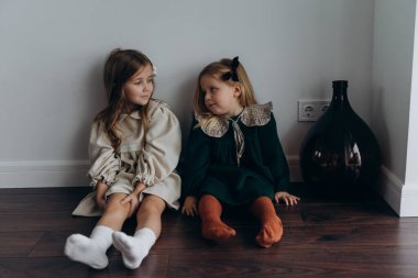 Güzel bohem elbiseler giymiş şirin küçük kızlar duvarın yanındaki ahşap zeminde birlikte oturuyorlar. 