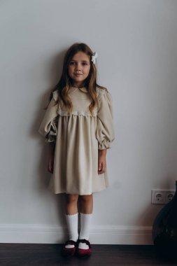 Güzel bir bohem elbisesi giymiş, duvarın yanındaki tahta zeminde poz vermiş, ayakta duran ve kameraya bakan küçük şirin bir kız. 