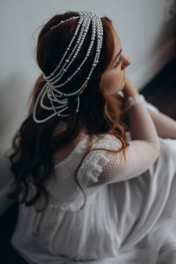 Beyaz bohem bir elbise giymiş güzel kızıl saçlı kadın ve inciden yapılmış başlığıyla ahşap zeminde düşünceli bir şekilde oturuyor.  