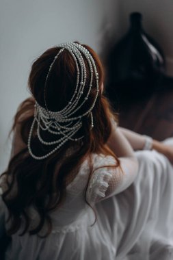 Beyaz bohem bir elbise giymiş güzel kızıl saçlı bir kadının arka manzarası ve ahşap zeminde düşünceli bir şekilde oturan inciden yapılmış bir başlığı.  