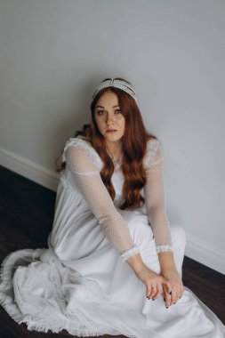 Beyaz bohem bir elbise giymiş güzel kızıl saçlı kadın ve inciden yapılmış başlığıyla ahşap zeminde düşünceli bir şekilde oturuyor.  