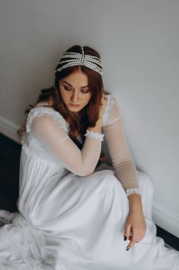 Beyaz bohem bir elbise giymiş güzel kızıl saçlı kadın ve inciden yapılmış başlığıyla ahşap zeminde düşünceli bir şekilde oturuyor.  