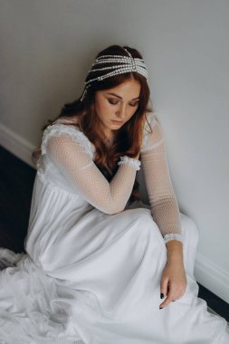 Beyaz bohem bir elbise giymiş güzel kızıl saçlı kadın ve inciden yapılmış başlığıyla ahşap zeminde düşünceli bir şekilde oturuyor.  