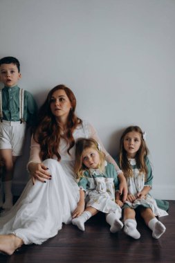 Kızları ve oğluyla birlikte beyaz bohem elbiseler giymiş güzel kızıl kadın köknar ağacının yanındaki tahta zeminde birlikte vakit geçiriyorlar. 