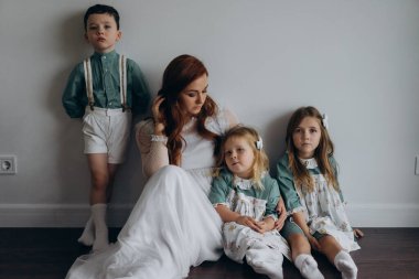 Kızları ve oğluyla birlikte beyaz bohem elbiseler giymiş güzel kızıl kadın köknar ağacının yanındaki tahta zeminde birlikte vakit geçiriyorlar. 