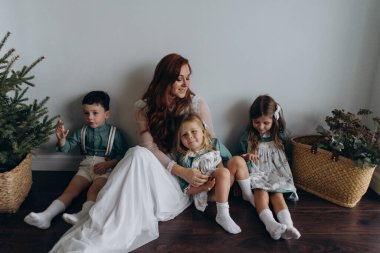 Kızları ve oğluyla birlikte beyaz bohem elbiseler giymiş güzel kızıl kadın köknar ağacının yanındaki tahta zeminde birlikte vakit geçiriyorlar. 