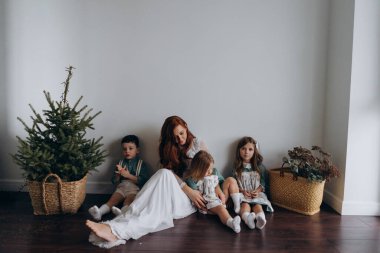 Kızları ve oğluyla birlikte beyaz bohem elbiseler giymiş güzel kızıl kadın köknar ağacının yanındaki tahta zeminde birlikte vakit geçiriyorlar. 