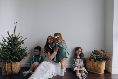 Kızları ve oğluyla birlikte beyaz bohem elbiseler giymiş güzel kızıl kadın köknar ağacının yanındaki tahta zeminde birlikte vakit geçiriyorlar. 