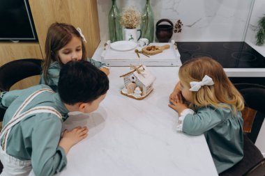 Çocuklar mutfaktaki masada zencefilli kurabiye evini inceliyor. 