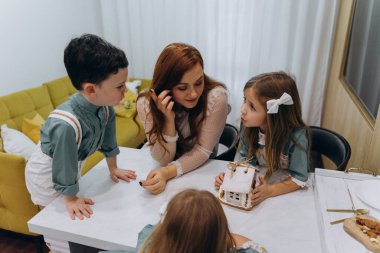 Beyaz bohem bir elbise giymiş güzel kızıl saçlı bir kadın küçük kızları ve oğluyla mutfakta zencefilli kurabiye yiyor.  