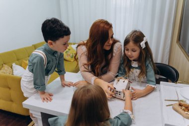 Beyaz bohem bir elbise giymiş güzel kızıl saçlı bir kadın küçük kızları ve oğluyla mutfakta zencefilli kurabiye yiyor. 