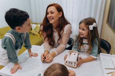 Beyaz bohem bir elbise giymiş güzel kızıl saçlı bir kadın küçük kızları ve oğluyla mutfakta zencefilli kurabiye yiyor.  