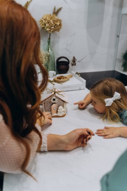 Beyaz bohem elbiseli güzel kızıl saçlı kadın ve küçük kızları mutfakta zencefilli kurabiye evini inceliyor. 