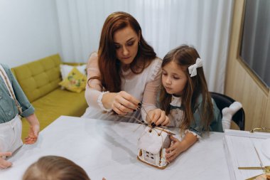 Kızları ve oğluyla birlikte beyaz bohem bir elbise giymiş güzel kızıl saçlı kadın mutfakta zencefilli kurabiye paketlerini açıyor. 