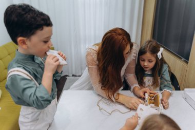 Beyaz bohem bir elbise giymiş güzel kızıl saçlı bir kadın küçük kızları ve oğluyla mutfakta zencefilli kurabiye yiyor. 