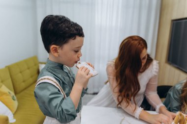 Beyaz bohem bir elbise giymiş güzel kızıl saçlı bir kadın küçük kızları ve oğluyla mutfakta zencefilli kurabiye yiyor.  