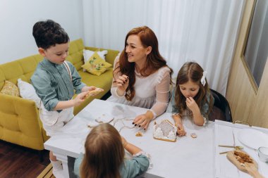 Güzel kızıl saçlı kadın beyaz bir bohem elbiseyle mutfakta küçük kızları ve oğluyla birlikte kurabiyeleri sütle yiyor.