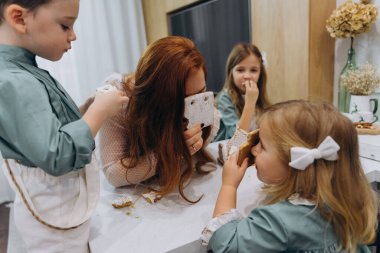 Güzel kızıl saçlı kadın beyaz bir bohem elbiseyle mutfakta küçük kızları ve oğluyla birlikte kurabiyeleri sütle yiyor.