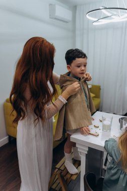 Beyaz bohem elbiseli güzel kızıl saçlı kadın mutfakta kurabiyeleri sütle yerken küçük oğluna bakıyor.