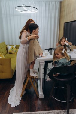 Güzel kızıl saçlı kadın beyaz bir bohem elbiseyle mutfakta küçük kızları ve oğluyla birlikte kurabiyeleri sütle yiyor.