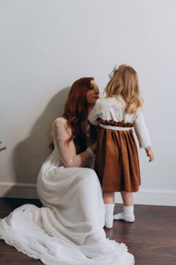 Beyaz bohem elbiseli güzel bir kadın küçük kızıyla köknar ağacının yanındaki ahşap zeminde oynuyor. 