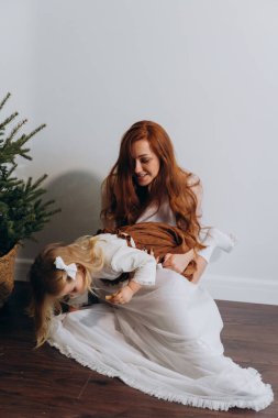 Beyaz bohem elbiseli güzel bir kadın küçük kızıyla köknar ağacının yanındaki ahşap zeminde oynuyor. 