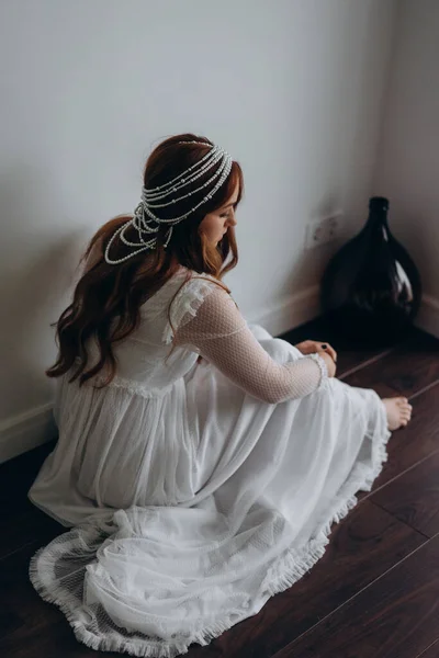 beautiful redhead woman dressed in a white bohemian dress and with a headdress made of pearl sitting thoughtfully on the wooden floor