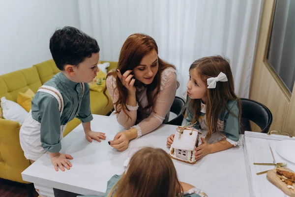 Beyaz bohem bir elbise giymiş güzel kızıl saçlı bir kadın küçük kızları ve oğluyla mutfakta zencefilli kurabiye yiyor.  