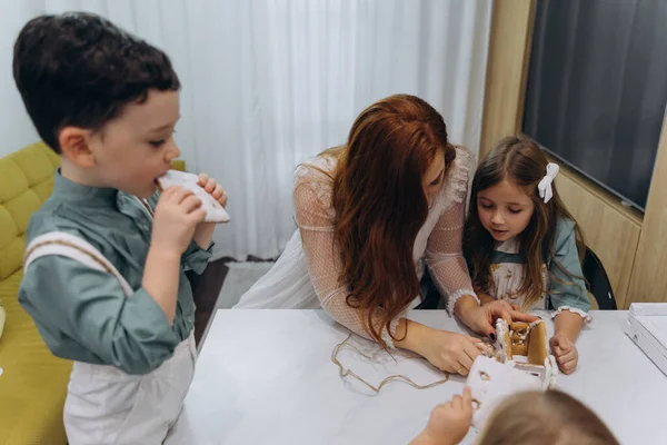 Beyaz bohem bir elbise giymiş güzel kızıl saçlı bir kadın küçük kızları ve oğluyla mutfakta zencefilli kurabiye yiyor. 
