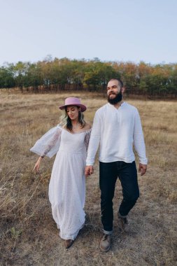 Kırsalda güzel bir çift, yakışıklı sakallı adam ve beyaz elbiseli kadın el ele yürüyorlar.