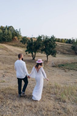 Kırsalda güzel bir çift, beyaz elbiseli yakışıklı sakallı adam ve kadın el ele tutuşuyorlar.