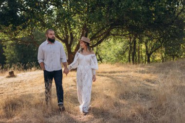 Kırsalda güzel bir çift, beyaz elbiseli yakışıklı sakallı adam ve kadın el ele tutuşuyorlar.