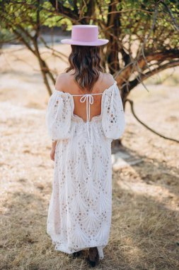 Arkadan görünüşlü, dövmeli, beyaz, uzun bir boho tarzı elbise ve kırsalda pembe şapkalı güzel bir kadın. 