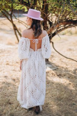 Arkadan görünüşlü, dövmeli, beyaz, uzun bir boho tarzı elbise ve kırsalda pembe şapkalı güzel bir kadın. 